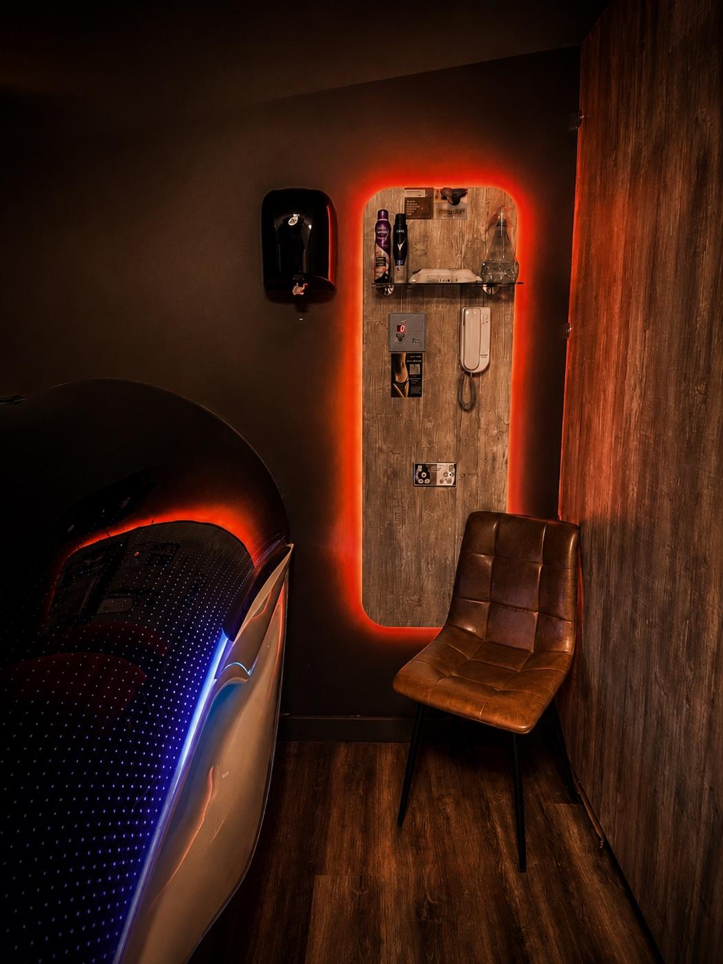 Another private suite at Tanners Lane Corby — a lounger lit in cool blue beside an amber-backlit amenity board and a leather chair