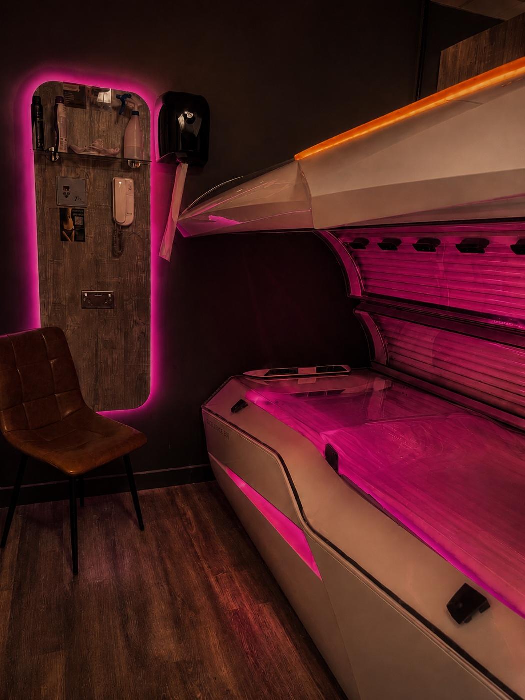 A private tanning suite at Tanners Lane Corby — a megaSun bed washed in warm magenta light, with a leather chair and a wood-panelled amenity board